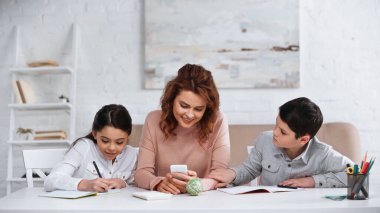Akıllı telefonlu gülümseyen anne oturma odasında çocuklarla ödev yapıyor. 