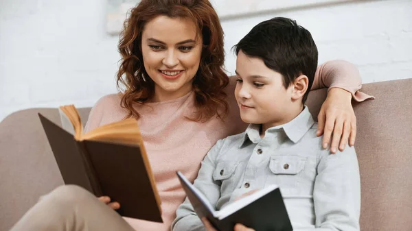 Mujer Sonriente Sosteniendo Libro Cerca Hijo Con Cuaderno Sofá Casa — Foto de Stock