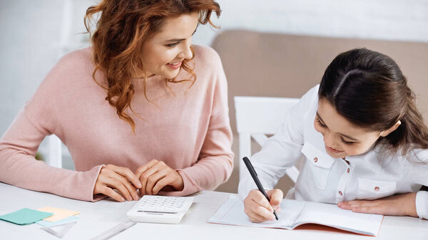Smiling woman looking at girl writing on notebook during education at home 