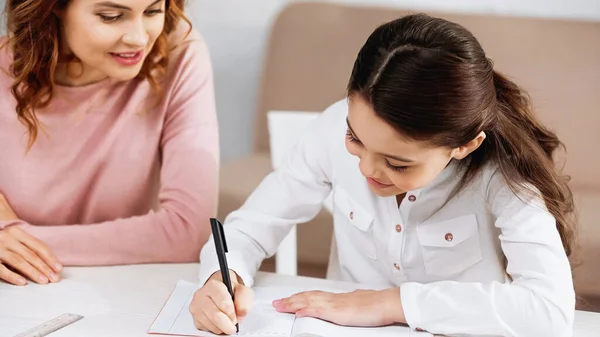 Criança Positiva Escrevendo Caderno Perto Mãe Fundo Embaçado Casa — Fotografia de Stock