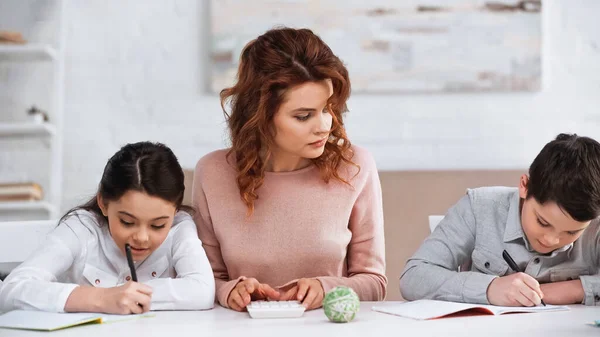 Vrouw Met Rekenmachine Buurt Van Kinderen Die Thuis Schriften Schrijven — Stockfoto