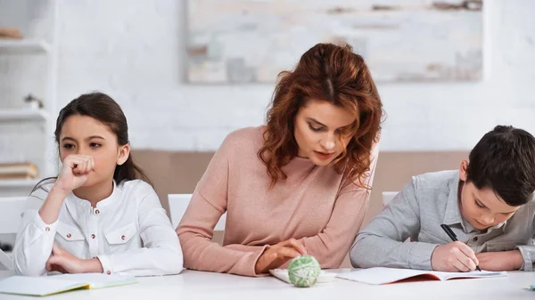 Woman using calculator near pensive daughter and son writing on notebook at home