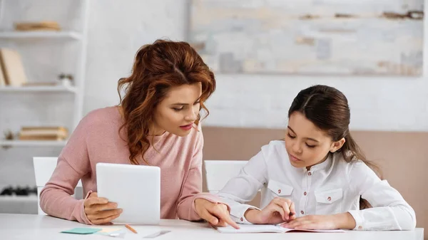 Mujer Con Tableta Digital Apuntando Cuaderno Cerca Hija Haciendo Tarea —  Fotos de Stock