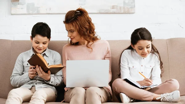 Sorrindo Menino Leitura Livro Perto Mãe Com Laptop Irmã Sofá — Fotografia de Stock