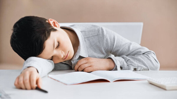 Boy with pen on blurred foreground lying near notebook and calculator on table 