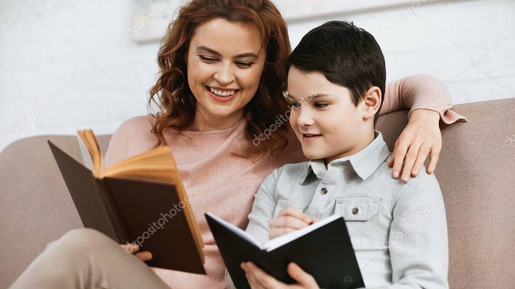 Cheerful woman reading book while helping son with homework 