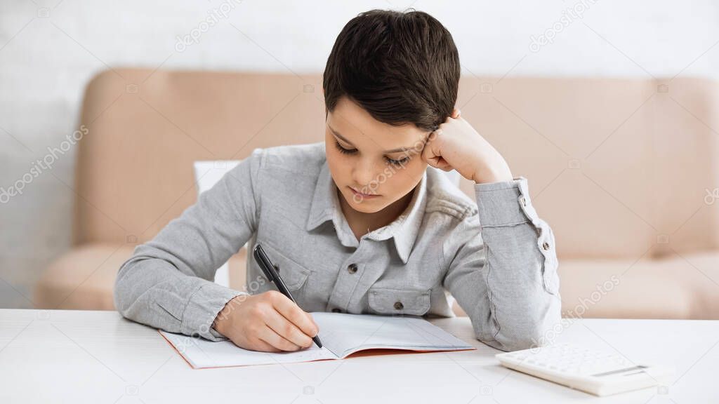 Boy writing on notebook near calculator on table at home 