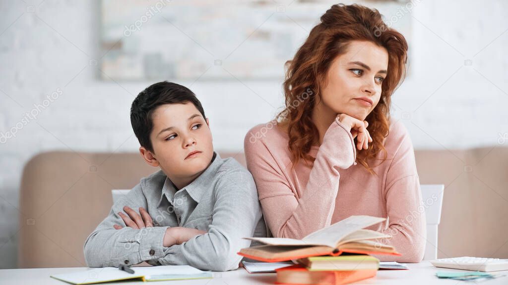 Displeased woman and son sitting near notebook and books at home 