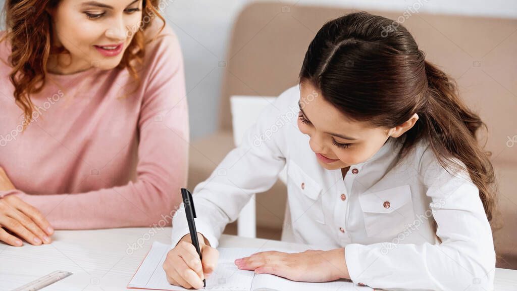 Positive kid writing on notebook near mother on blurred background at home 
