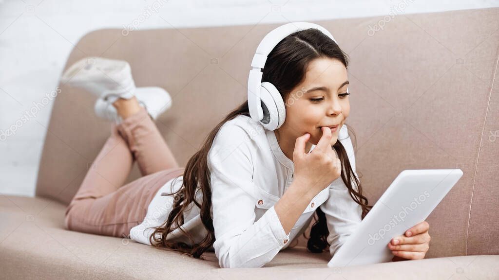 Smiling kid using headphones and digital tablet on couch in living room 