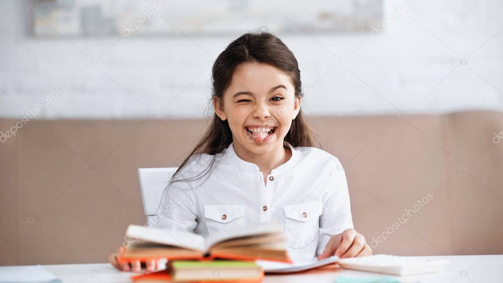 Preteen kid sticking out tongue near books on blurred foreground 