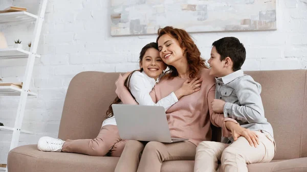 Donna Che Abbraccia Bambini Sorridenti Vicino Computer Portatile Sul Divano — Foto Stock