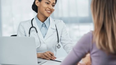 Beyaz önlüklü mutlu Afro-Amerikan doktor masadaki bilgisayarın yanında bulanık hastayla konuşuyor.