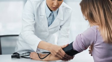 Beyaz önlüklü Afrikalı Amerikalı doktorun klinikteki kadının kan basıncını ölçtüğü görüntüler.