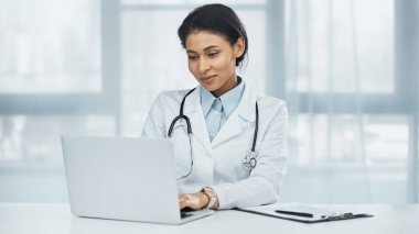 Sırıtan Afrikalı Amerikalı doktor masadaki panonun yanında dizüstü bilgisayar kullanıyor.