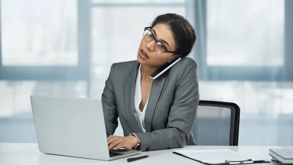 非洲裔美国女商人 戴着眼镜 在桌面上笔记本电脑旁边用智能手机交谈 — 图库照片