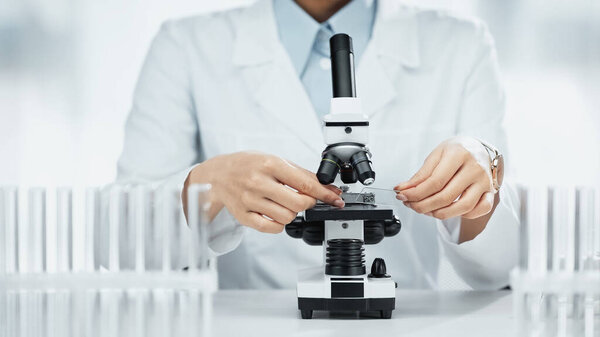 cropped view of african american scientist holding glass with sample near microscope in lab 