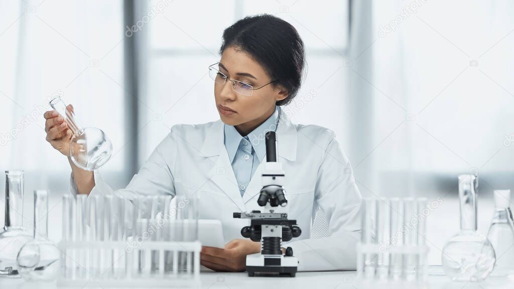 african american scientist in glasses holding flask near microscope 