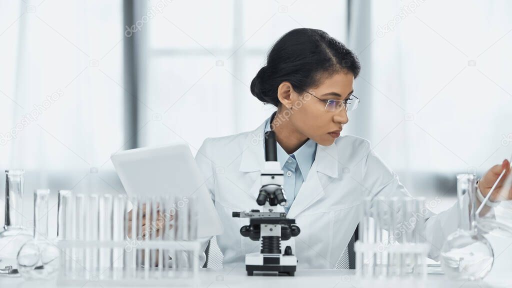 african american scientist in glasses holding digital tablet near microscope and test tubes in lab 