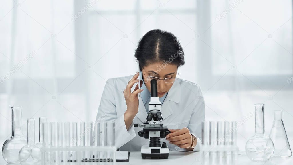 african american scientist in glasses talking on smartphone while looking through microscope in lab 