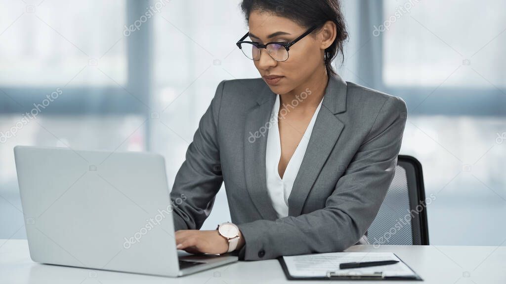 african american businesswoman in glasses using laptop near clipboard on desk