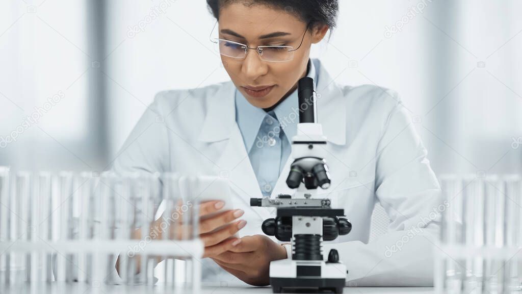 african american scientist looking and smartphone near microscope in lab