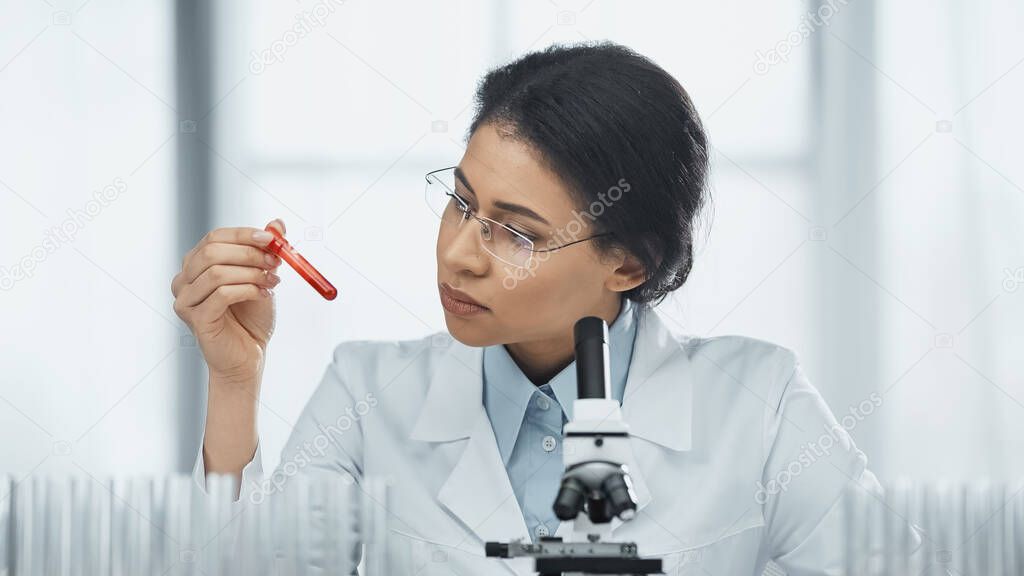 african american scientist in glasses holding test tube with blood sample near microscope in lab 