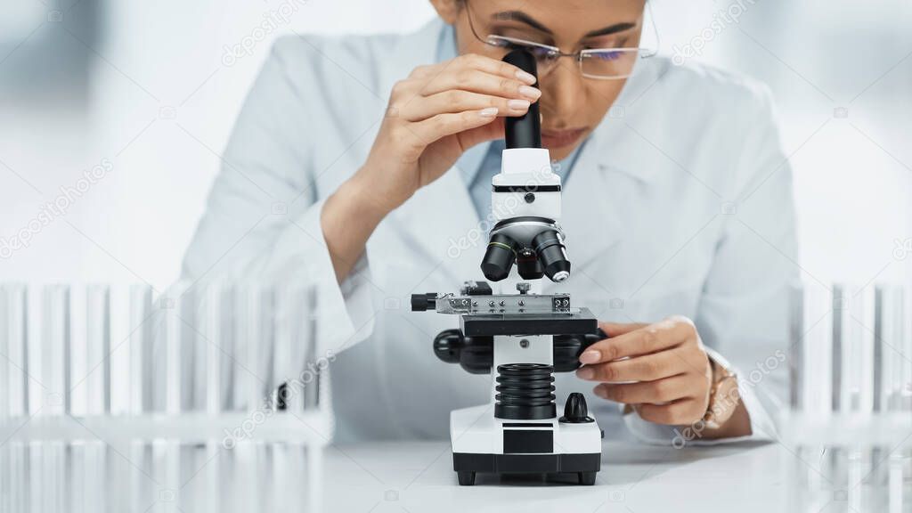 focused african american scientist looking through microscope near test tubes in lab 