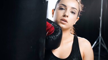Spor eldiveni giymiş yorgun sporcu kadın spor salonunda kum torbasına yaslanıyor. 