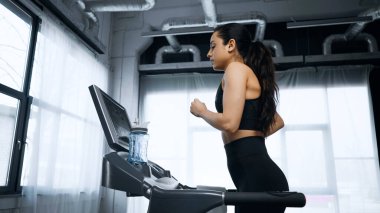 side view of sportswoman running on treadmill near sports bottle with water in gym clipart