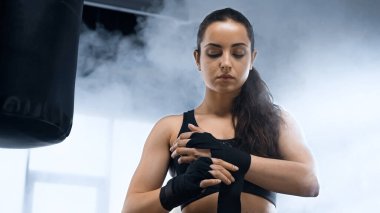 Genç esmer sporcu kadın yumruk torbasının yanında ellerini bandajlıyor. 