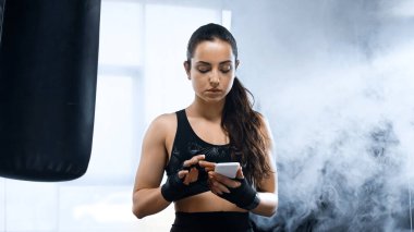 Genç esmer sporcu kadın ellerinde boks bandajlarıyla akıllı telefon kullanıyor. 