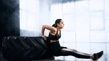 Esmer ve sportif genç bir kadın spor salonunda lastiğin yanında egzersiz yapıyor. 
