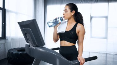 Spor kadını koşu bandında yürüyor ve spor salonunda su içiyor.