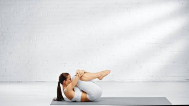 Beyaz spor giyim giymiş esmer bir kadının evde yoga çalışmasının yan görüntüsü. 