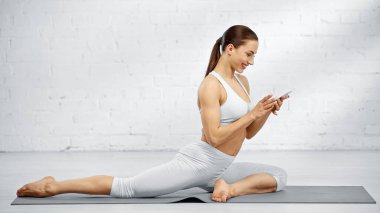 Neşeli sporcu kadın yoga minderinde akıllı telefon kullanıyor. 