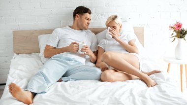 Gülümseyen genç adam kız arkadaşına bakıyor yatakta kahve içiyor.