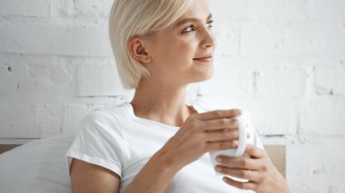 Beyaz tişörtlü gülümseyen kadın yatak odasında bir fincan kahve tutuyor.