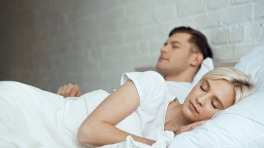 Sarışın kadın, arka planda bulanık bir adamla yatakta uyuyor. 