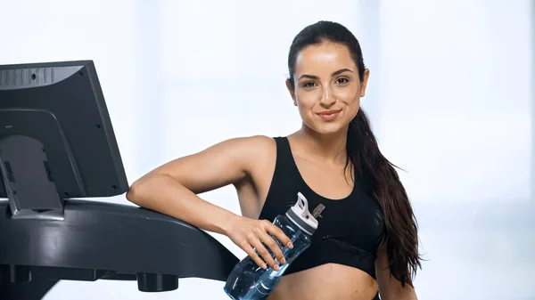 穿着运动服的年轻而快乐的女人在跑步机附近拿着装有水的运动瓶子 — 图库照片