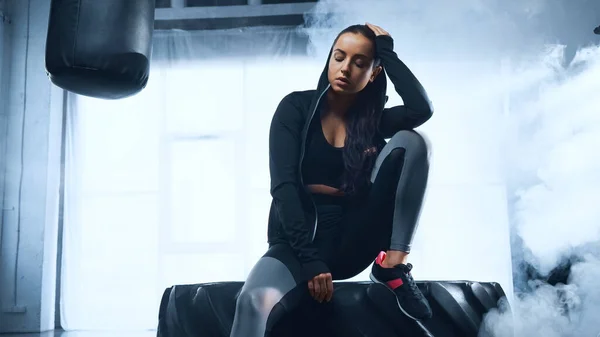 sporty woman in hoodie sitting on tire near punching back and smoke behind