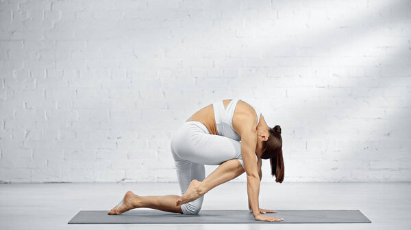 Side view of woman in white sportswear bending on yoga mat 