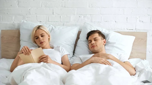 Young Blonde Woman Reading Book Sleeping Boyfriend Bedroom — Stock Photo, Image