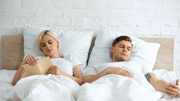 Young Blonde Woman Reading Book Boyfriend Using Smartphone Bedroom — Stock Photo, Image