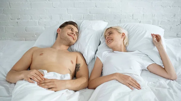 happy tattooed man and blonde woman looking at each other in bed