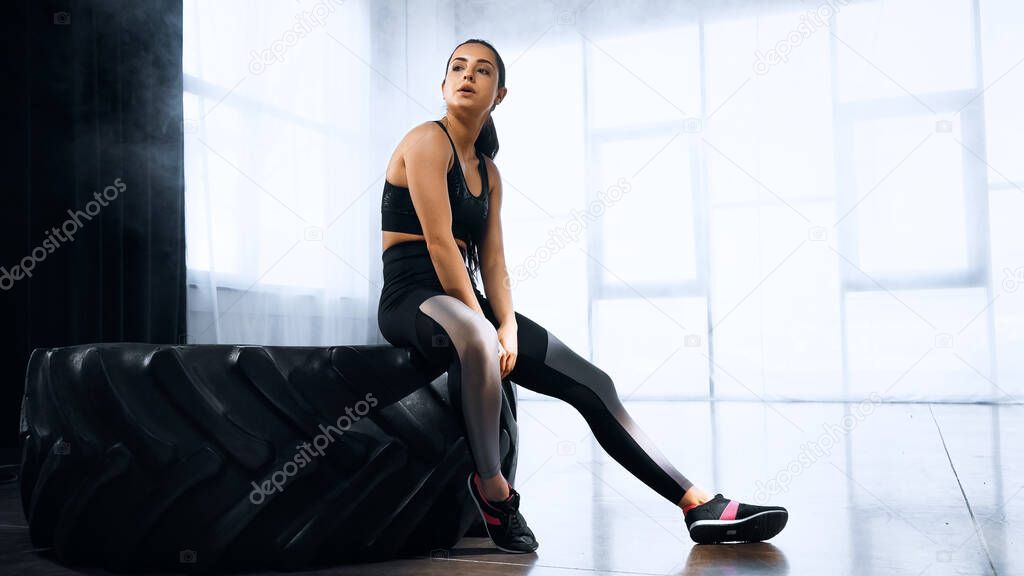 tired young sportswoman sitting on tire while resting in sports center