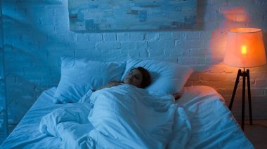 Woman sleeping on bed near floor lamp in bedroom  clipart