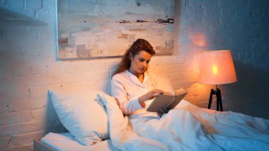 Pijamalı bir kadın, yatak odasındaki lambanın yanında kitap okuyor. 