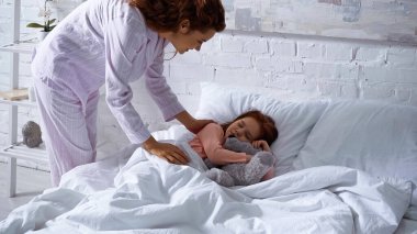 Pijamalı bir kadın. Çocuğun yanında duruyor. Yatakta uyuyor. 