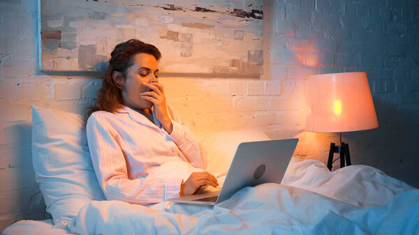 Женщина в пижаме зевает во время использования ноутбука на кровати вечером 